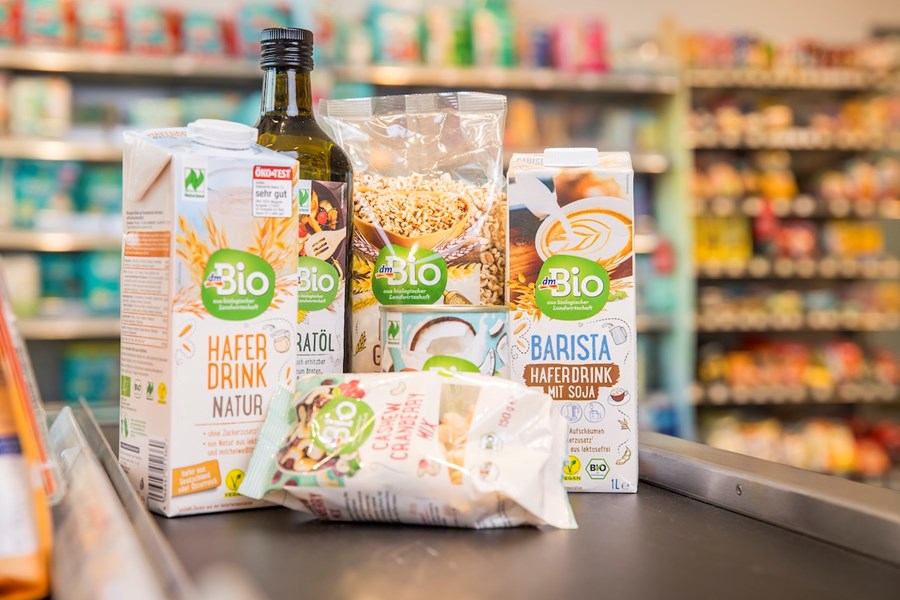 Mehrere dmBio-Produkte stehen auf einem Supermarktband: zwei Haferdrinks, ein Flasche Bratöl, eine Packung Getreide und ein Beutel Knuspermüsli. Im Hintergrund sind unscharf Regale mit weiteren Produkten zu sehen.
